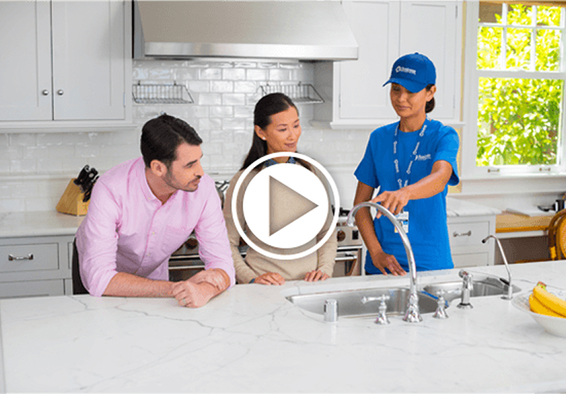 Video thumbnail of a home services technician in a blue uniform demonstrating a kitchen faucet to a seated couple at a white marble island in a bright, modern kitchen, with a large play button icon centered over the image.