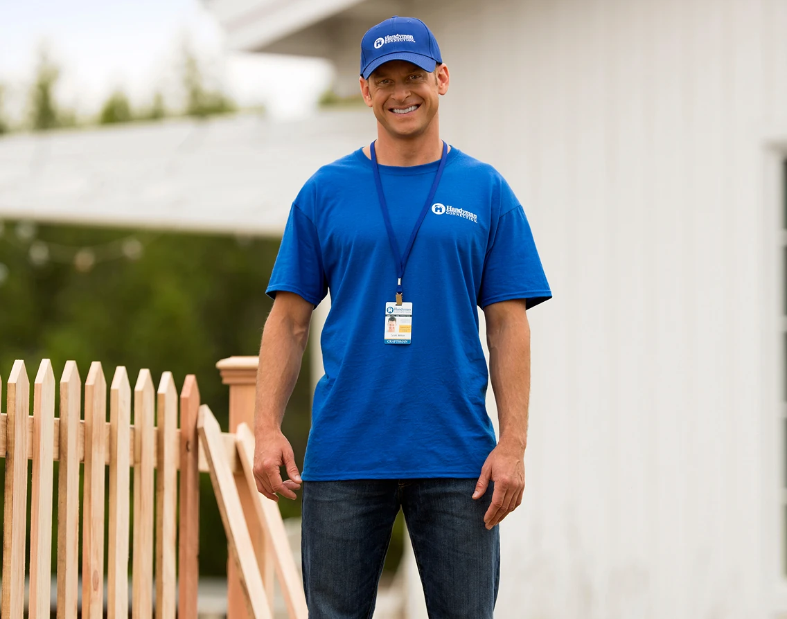 Smiling contractor in a blue cap and T-shirt with a name badge on a lanyard stands beside a new wooden picket fence, with a house blurred in the background.