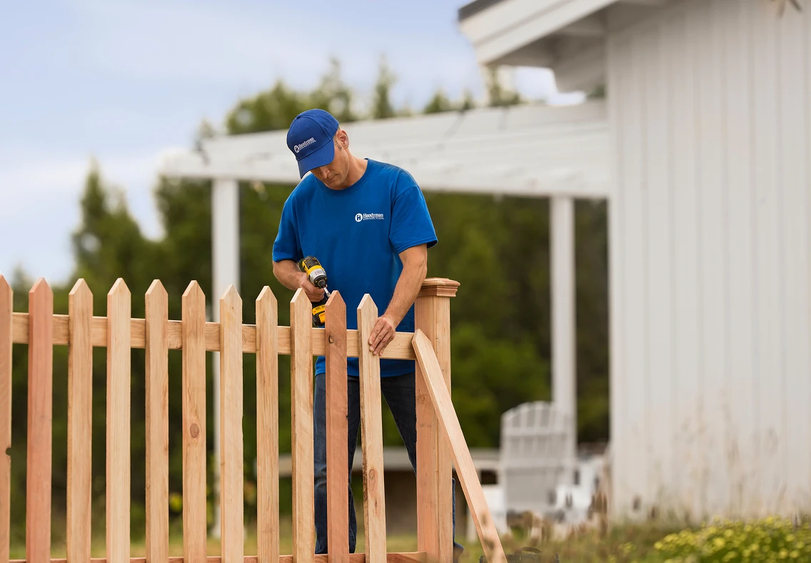 Contractor in a blue shirt and cap uses a power drill to attach pickets to a wooden fence gate, with a white house and pergola blurred in the background.