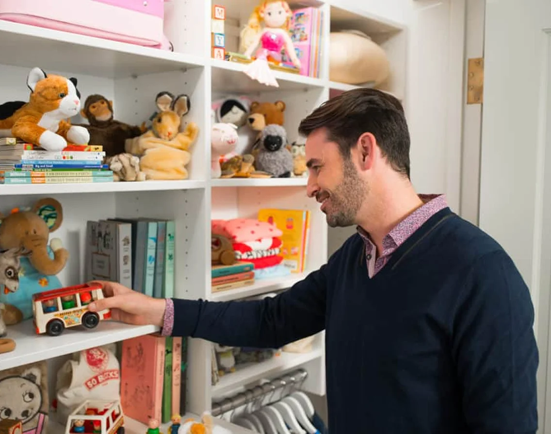 Man arranging children’s toys and books on built-in shelving in a child’s room or nursery.