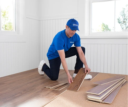Technician in a Handyman Connection uniform kneels on the floor positioning a wood-look plank, with additional flooring boards stacked nearby.