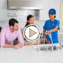 Video thumbnail of a home services technician in a blue uniform demonstrating a kitchen faucet to a seated couple at a white marble island in a bright, modern kitchen, with a large play button icon centered over the image.