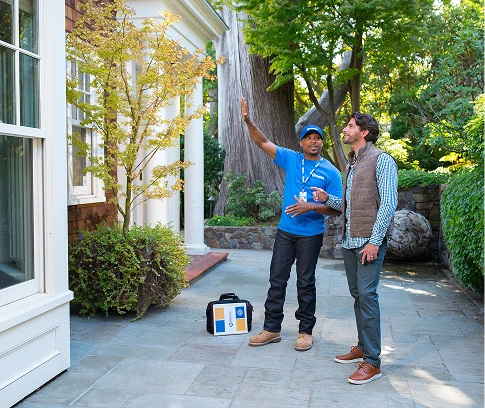 Handyman Connection technician in a blue uniform speaks with a homeowner on a stone patio, gesturing toward the exterior of the house and nearby trees as they discuss outdoor work.