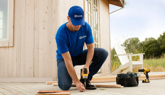 Home service professional kneeling on a deck using a power drill to secure wooden boards.