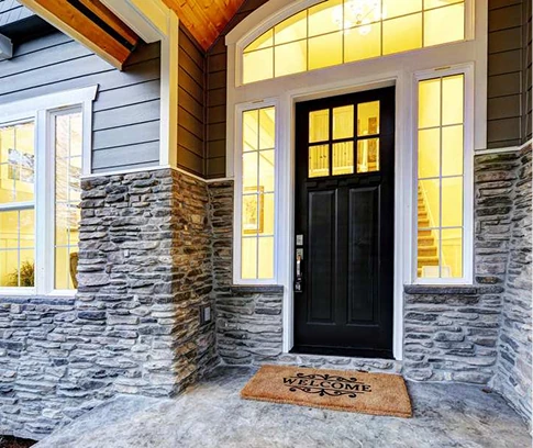 Front entry with a black door, sidelights, and a “Welcome” doormat beneath a covered porch with stone pillars.