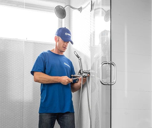 Technician in a blue Handyman Connection uniform stands in a tiled shower adjusting the shower fixture with a wrench beside a glass shower door.